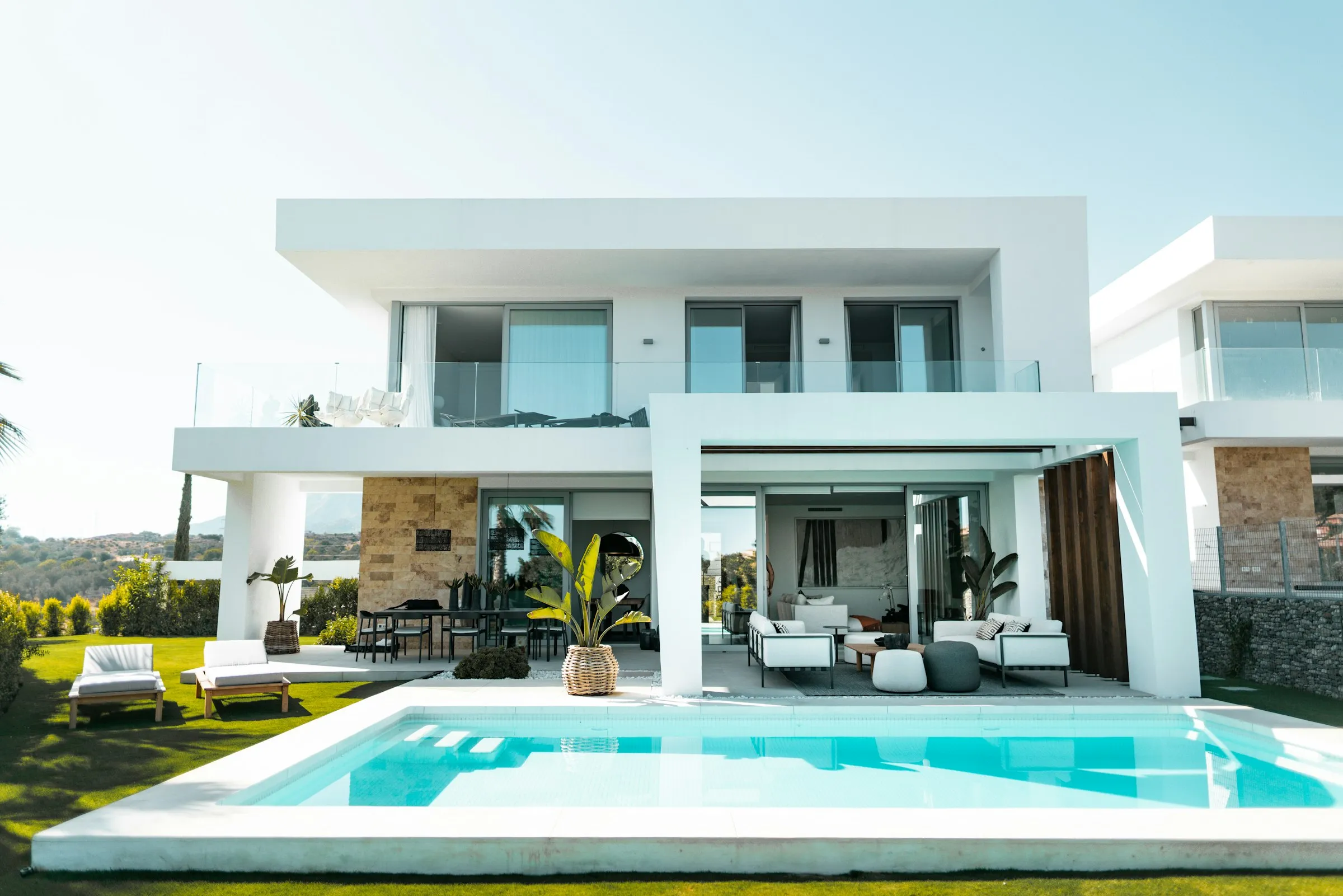 Villa blanca de una planta con piscina desbordante y palmeras, al sol de la tarde